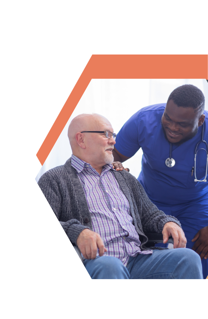 A smiling healthcare worker in blue scrubs and a stethoscope gently places a hand on the shoulder of an older man in a wheelchair, both sharing a friendly moment in a caring South Central Minnesota setting.