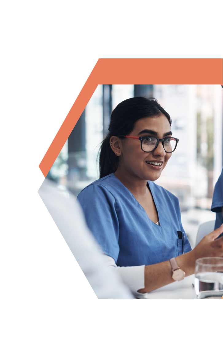 A smiling healthcare worker in blue scrubs and glasses sits at a table, holding a pen, with a glass of water nearby, framed by an orange hexagon—representing CNA Training in Central Minnesota.