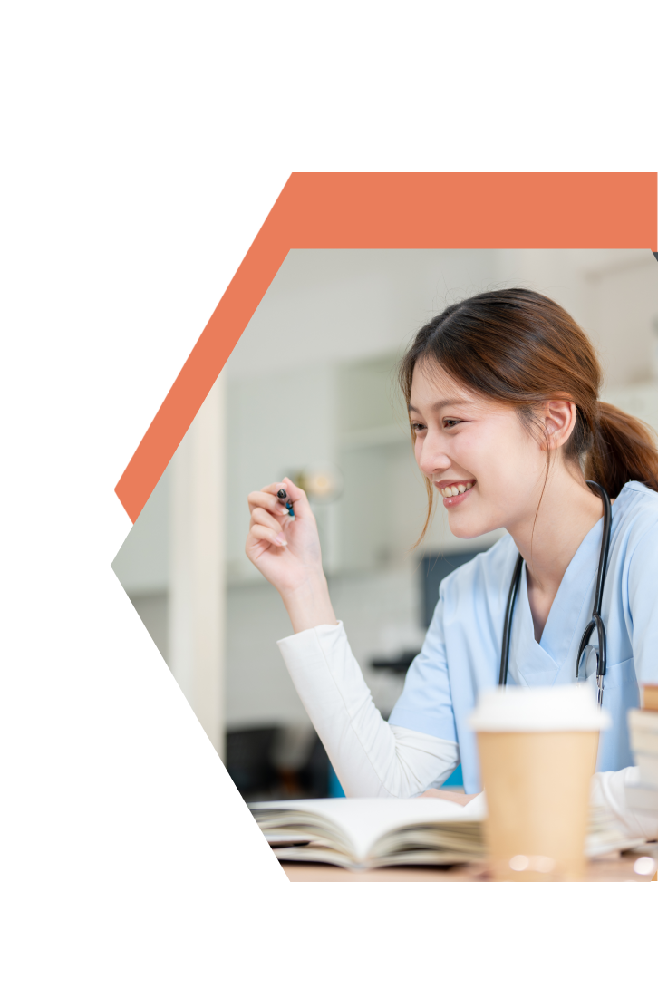 A smiling woman in medical scrubs with a stethoscope sits at a desk with an open book and coffee, holding a pen, appearing engaged and focused—ready to take on her CNA Training in Northwest Minnesota.