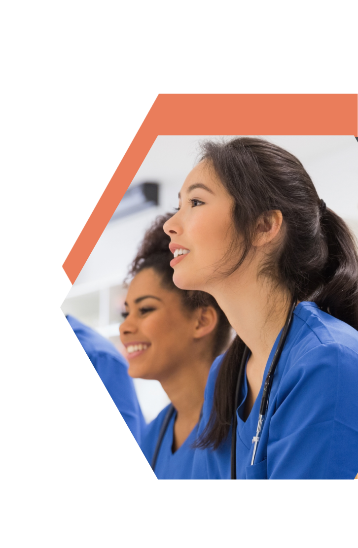 Two young women in blue medical scrubs, smiling and attentive, sit side by side—one with a stethoscope around her neck. The image, framed with a hexagonal orange border on white, reflects CNA Training in Central Lakes Minnesota.