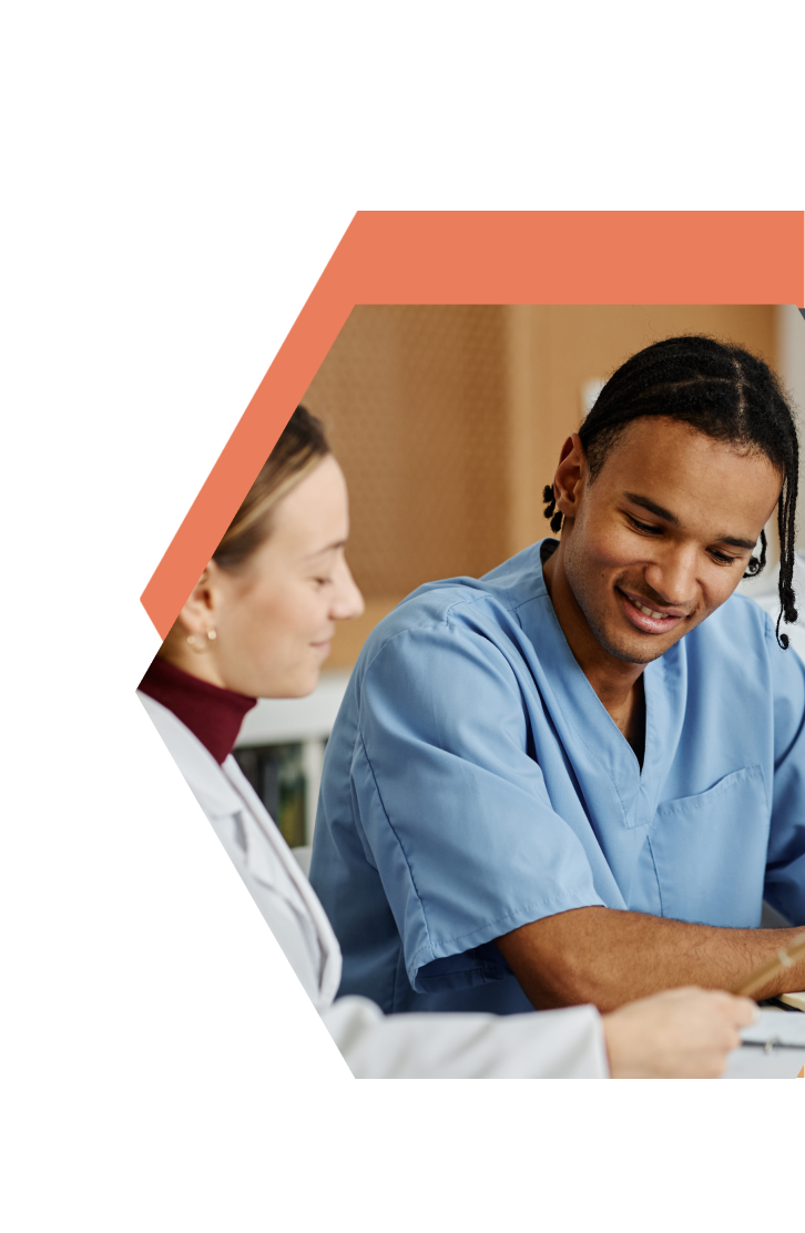 Two healthcare professionals, one in blue scrubs and the other in a white coat, sit together at a table, smiling and discussing CNA Training in East Central Minnesota in a bright, modern setting.